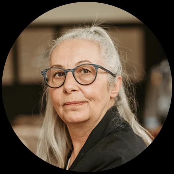 Elderly woman with long gray hair in a ponytail, wearing glasses and a black top, looking slightly to the side against a blurred background.